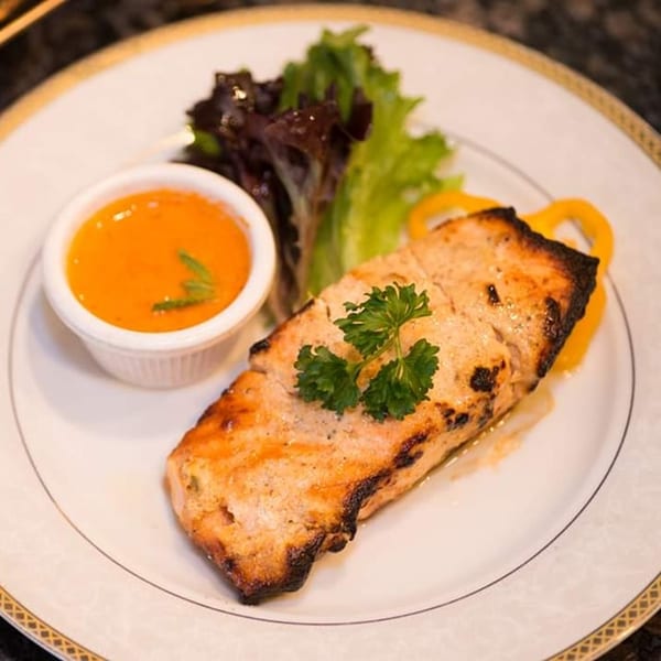 Grilled salmon with a side of orange sauce, fresh greens, and yellow bell pepper on a white plate.