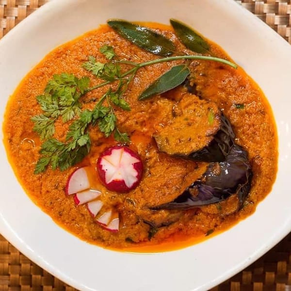 Indian eggplant curry with radishes and herbs in a white bowl on a woven mat.
